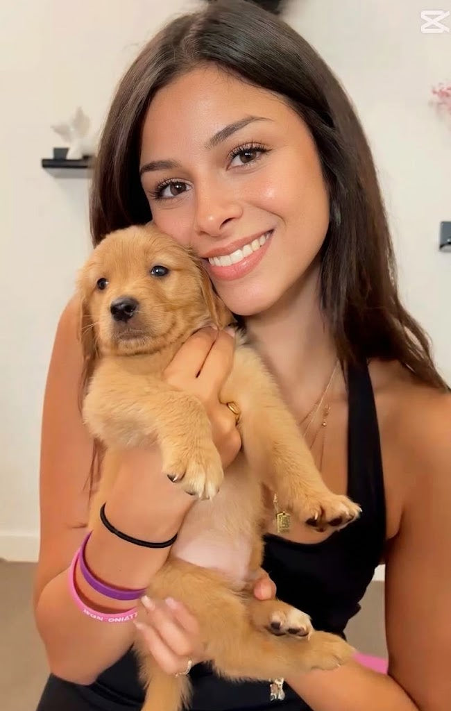 A brunette woman holds a small golden retriever puppy and smiles at the camera. They are sitting on the floor of a yoga studio.